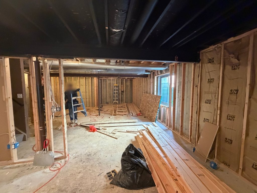 Unfinished basement during framing, with new wood stud walls, faced batt insulation on the exterior walls, a window, and stacked lumber on the concrete floor, with a worker near a ladder in the background