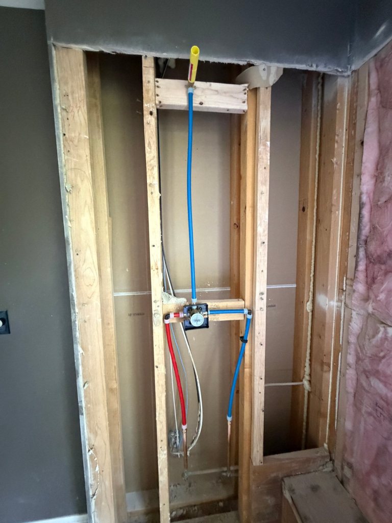Shower valve roughed in between wood studs, with blue and red PEX supply lines and a capped shower head riser, next to a wall of pink batt insulation