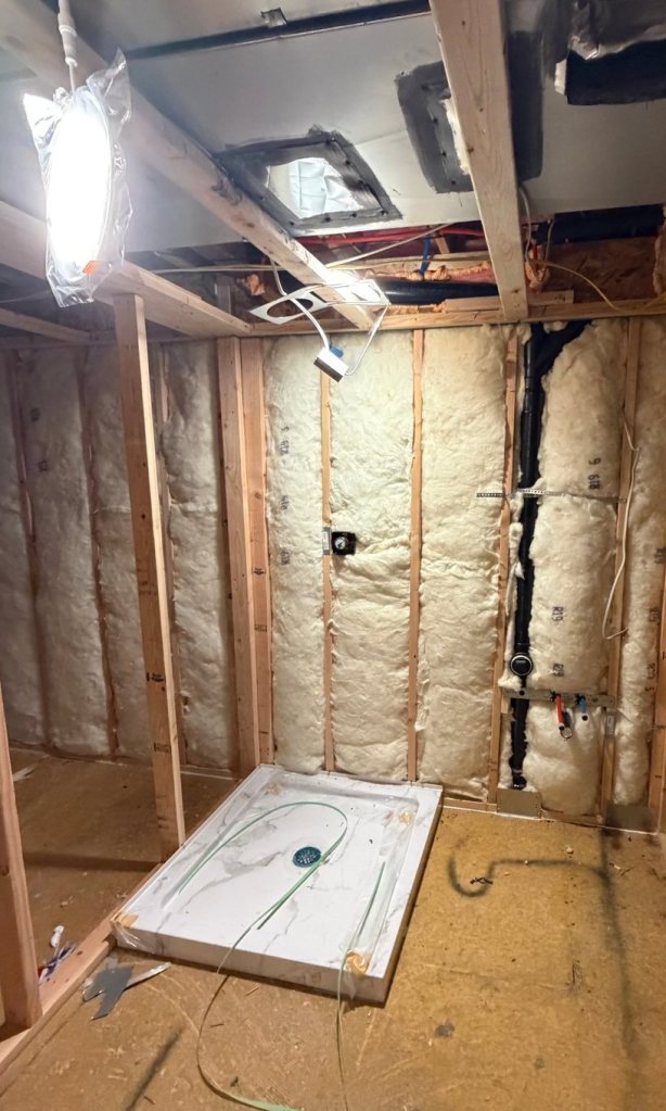 Room at rough-in with a white shower pan set on the subfloor, batt insulation in the stud walls, and a black drain line and supply stub-outs roughed in