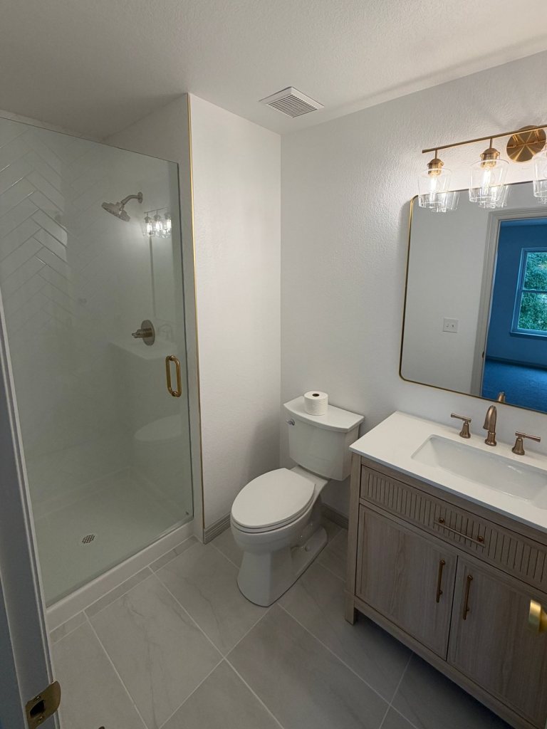 Finished bathroom with a glass shower enclosure over white herringbone tile, a wood vanity with a white top, gold faucet and light fixture, and a gold-framed mirror