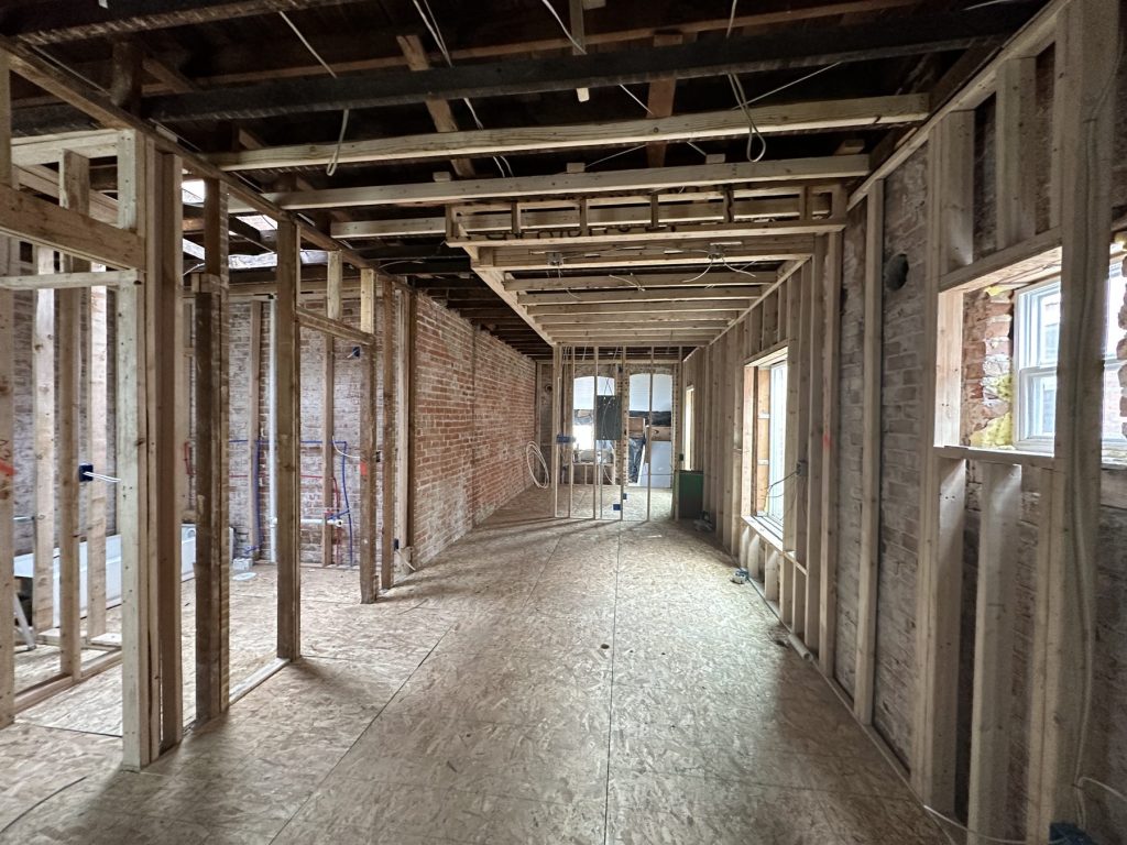 Open interior mid-remodel with new wood stud walls framed along exposed brick, open ceiling joists, and OSB subfloor