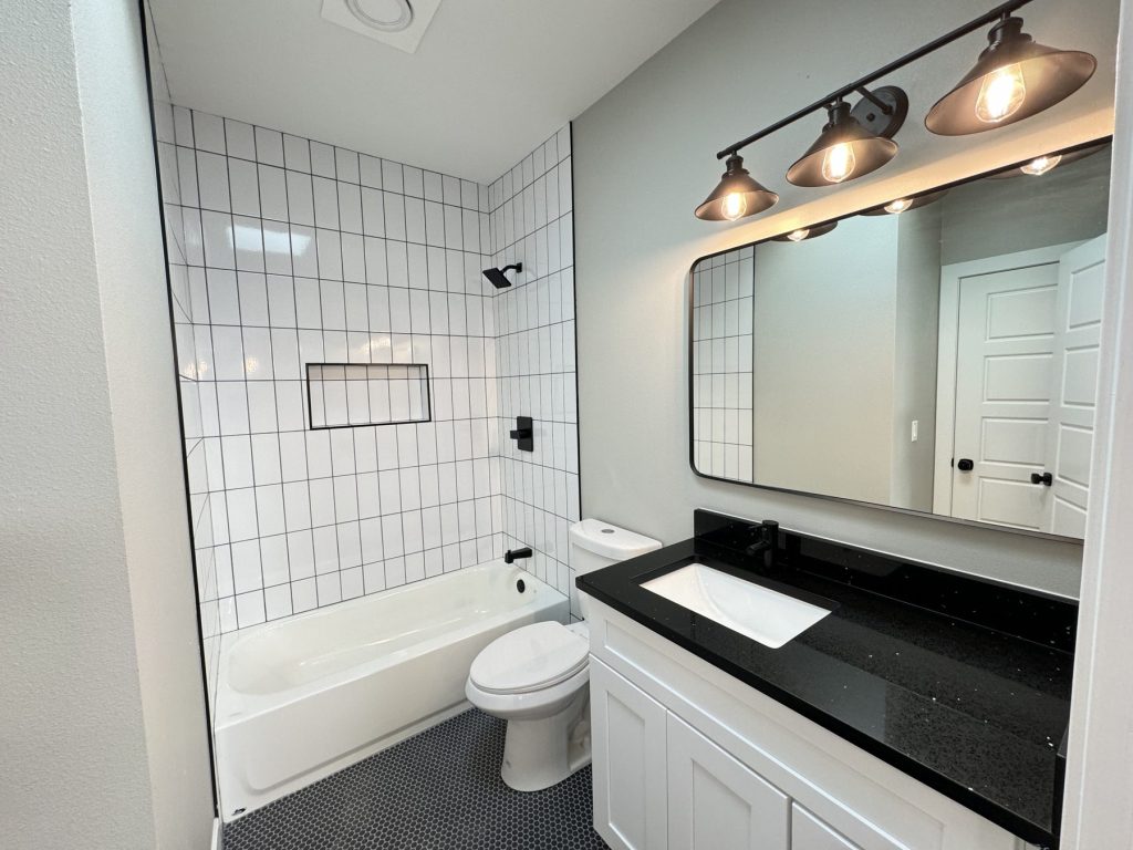 Remodeled bathroom with a white vanity and black quartz top, large mirror and black light bar with copper shades, tub with white vertical subway tile surround and niche, and black hex floor tile