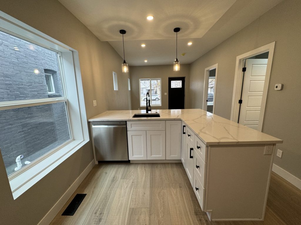 Remodeled kitchen with white shaker cabinets, a quartz-topped peninsula with undermount sink and black faucet, stainless dishwasher, pendant lights, and wood-look plank flooring