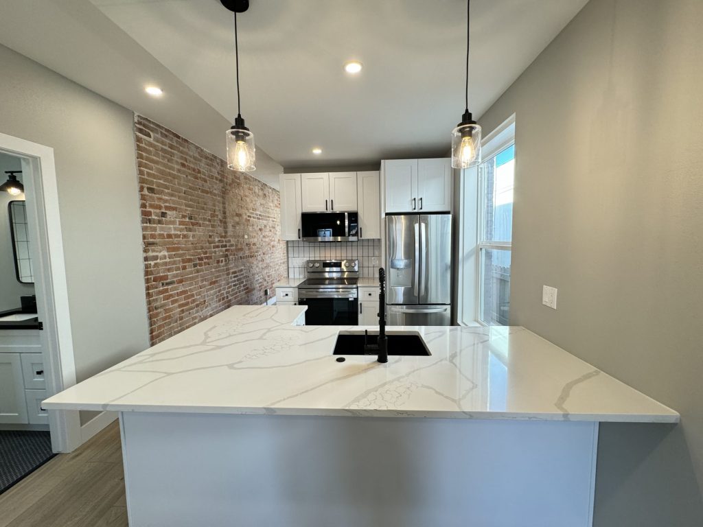 Remodeled kitchen seen straight on: large quartz peninsula with black undermount sink, white shaker cabinets, stainless range and refrigerator, subway tile backsplash, and an exposed brick wall
