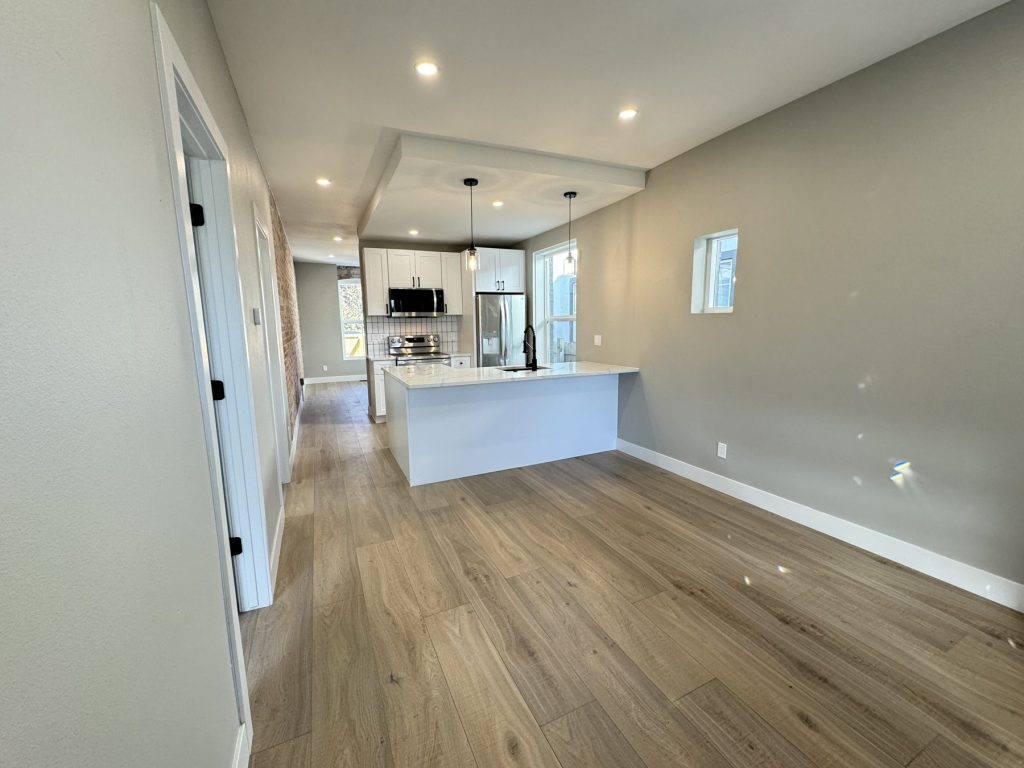 Open living area after remodeling with wood-look plank flooring, recessed lighting, gray walls, and a white kitchen with quartz peninsula and pendant lights beyond