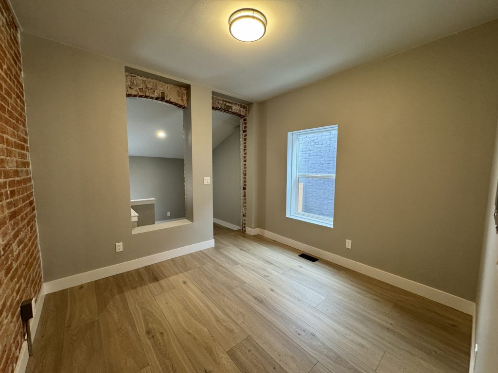 Finished room with two arched openings trimmed in original exposed brick, an exposed brick side wall, new window, wood-look plank flooring, and white baseboards