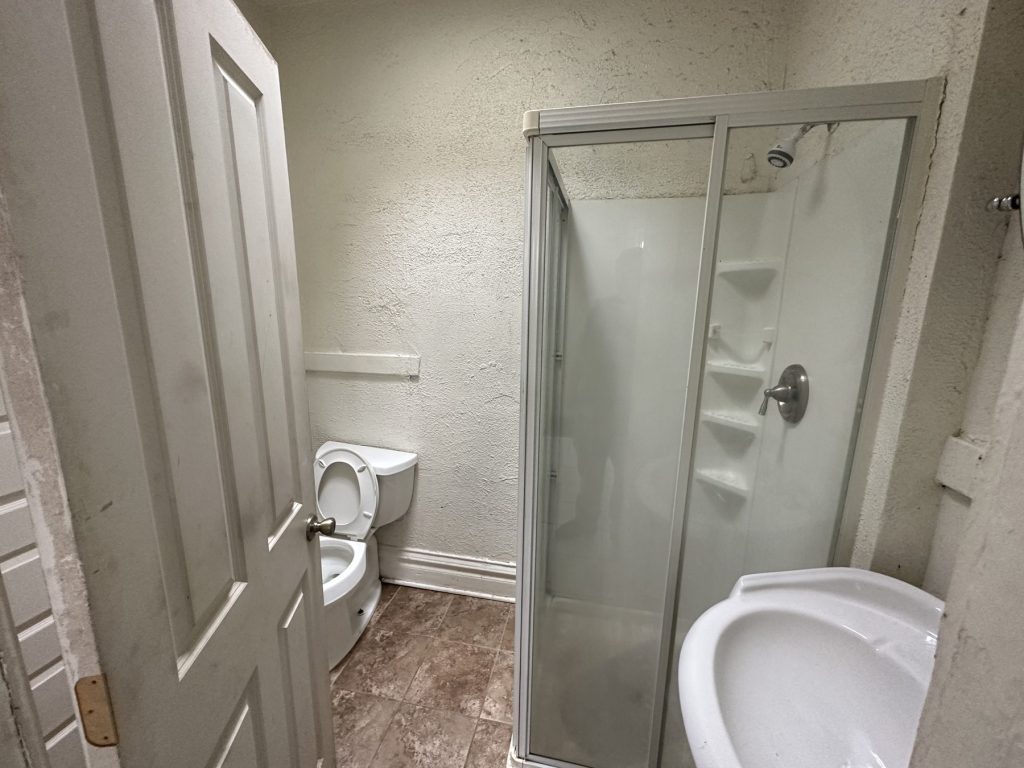 Small bathroom before remodeling with a corner shower stall, pedestal sink, textured walls, and brown vinyl floor tile