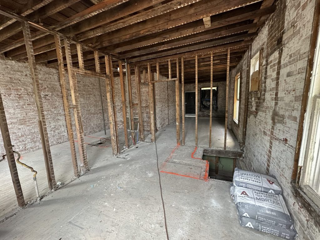 Interior fully gutted to the brick walls, old studs and ceiling joists exposed, layout marked in orange paint on the subfloor, and bags of mortar staged by the wall