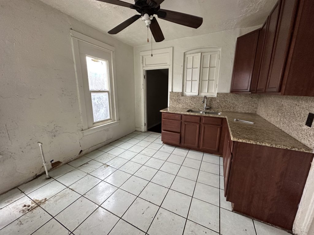 Kitchen before remodeling with dated cherry cabinets, granite counters, worn white floor tile, a ceiling fan, and damaged plaster walls