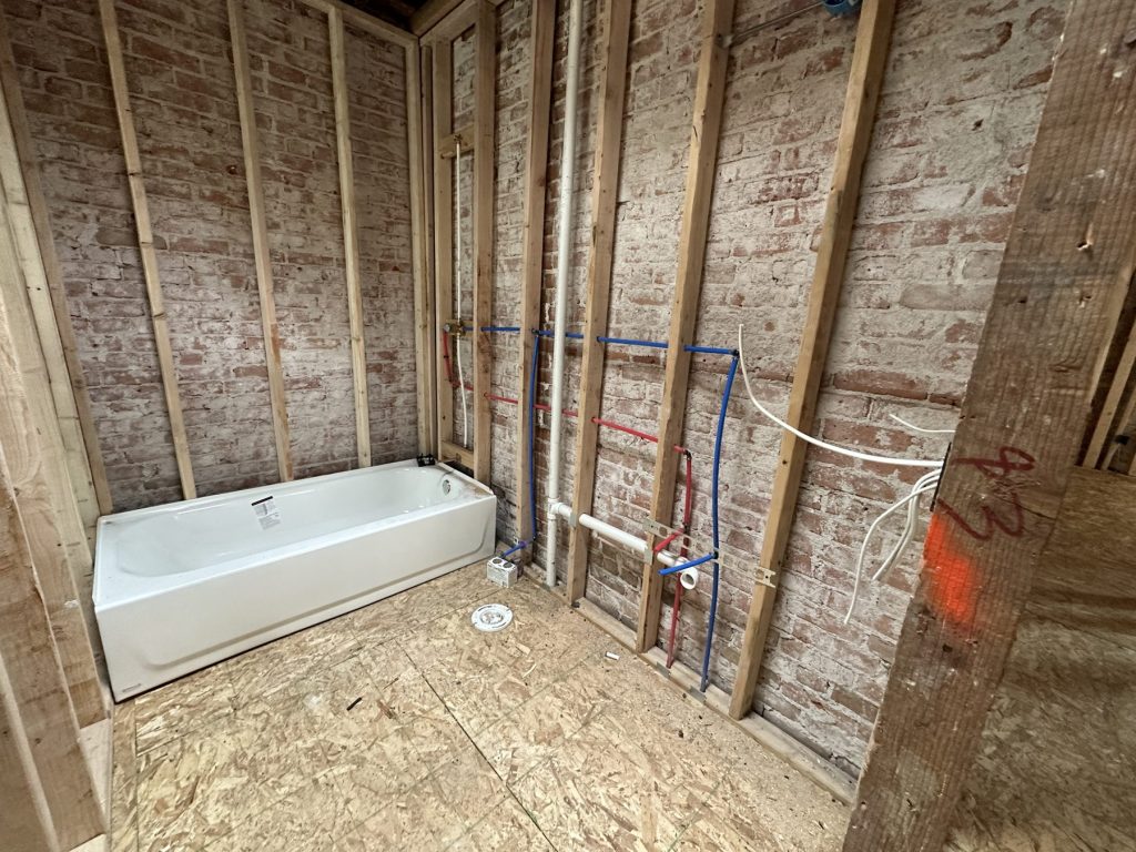 Bathroom under construction with a new white bathtub set, wood stud framing over exposed brick, and red and blue PEX supply lines and PVC drain roughed in