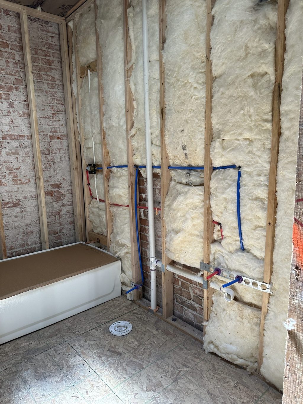 Bathroom wall framing filled with batt insulation, with PEX water lines, shower valve, and PVC drain and vent roughed in beside a protected bathtub