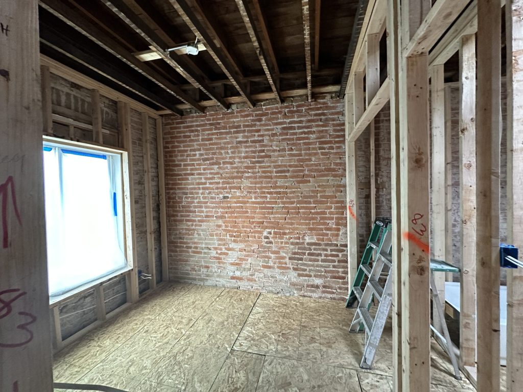 Bedroom mid-remodel with new stud framing around a window, an exposed red brick wall, open ceiling joists, and a new OSB subfloor