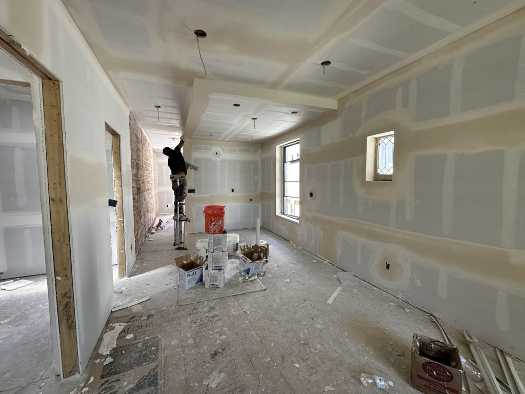 Drywall taping and mudding in progress in a long room with a tray ceiling with soffits, recessed light openings, and an exposed brick wall at the far end