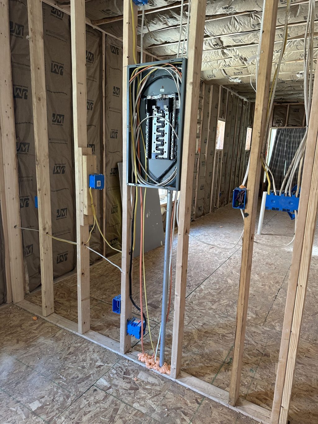 New electrical panel mounted between wall studs with circuits roughed in, blue outlet boxes on the framing, and insulated exterior walls
