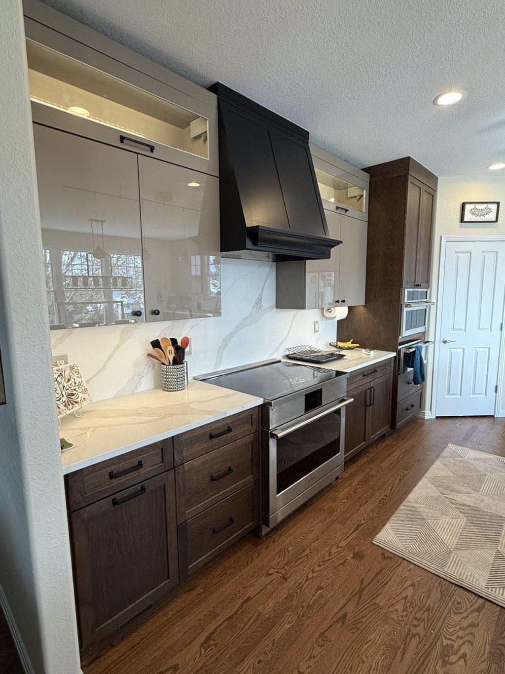 Remodeled kitchen with dark wood lower cabinets, glossy upper cabinets with lit glass tops, a black range hood, a full-height marble-look quartz backsplash, and a stainless range
