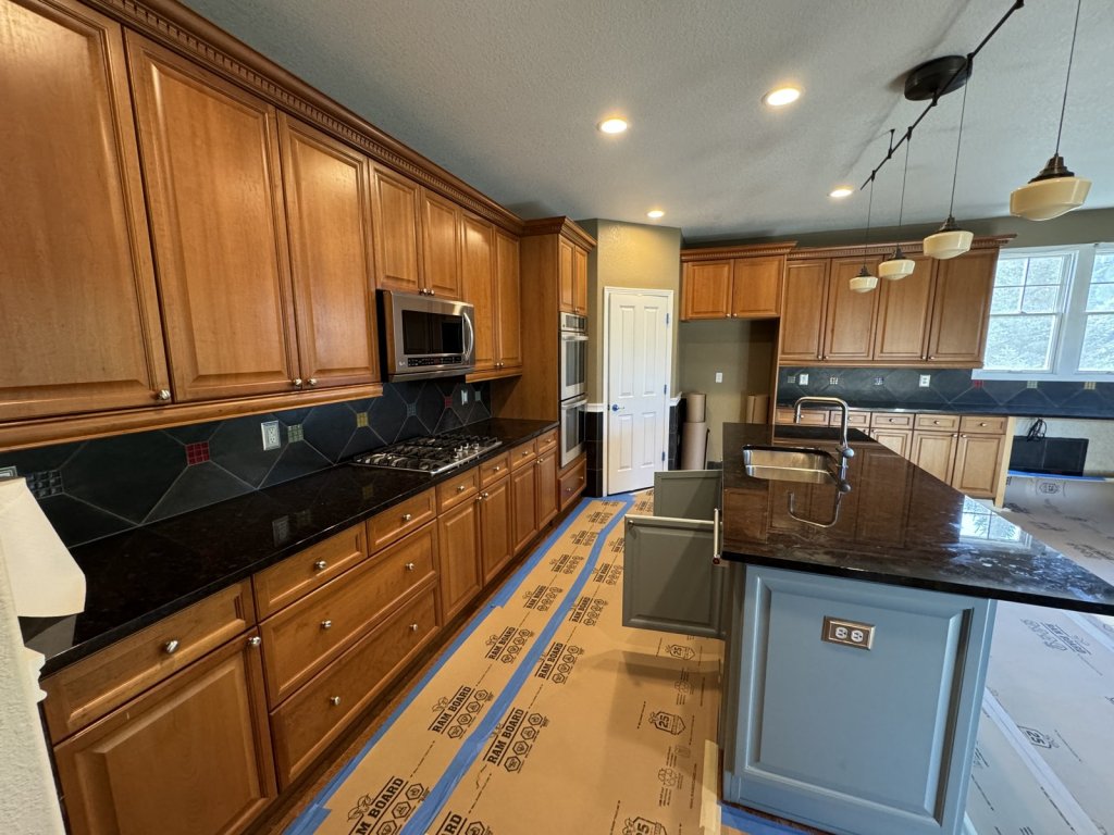 Kitchen before remodeling with honey oak cabinets, black granite counters, a dark tile backsplash, and a gray-blue island, floors covered in protective board