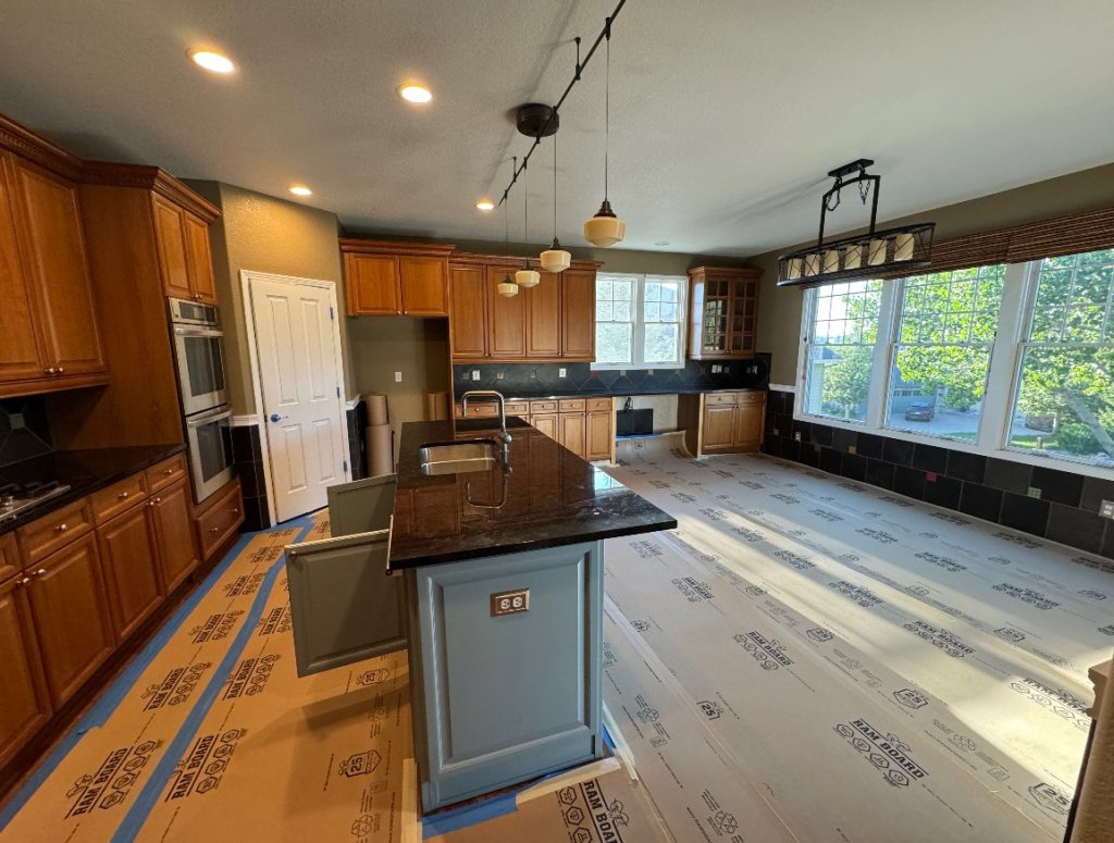 Wide view of the kitchen before remodeling: oak cabinets, a granite-topped island, pendant track lights, and a bank of windows over a dark tile wainscot