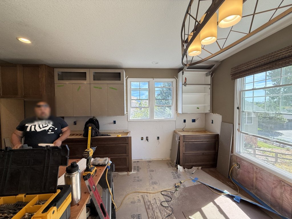 Kitchen mid-remodel with new dark wood base cabinets and light gloss upper cabinets being installed, new outlets on bare drywall, and tools on a work table