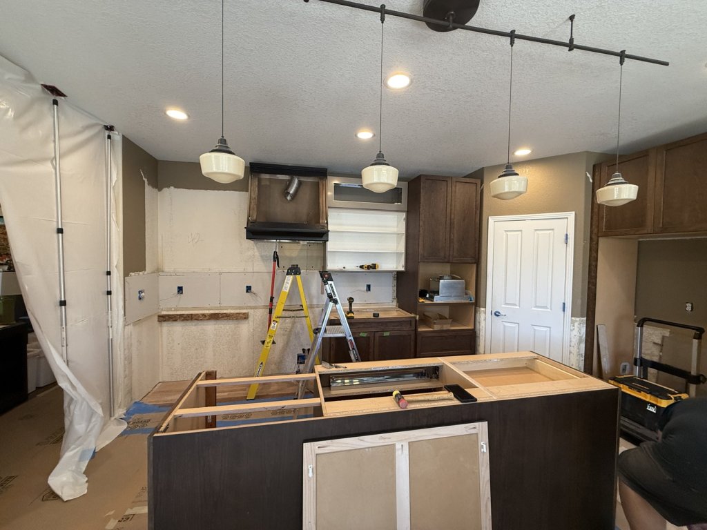 Kitchen mid-remodel: new island cabinets set and framed for a cooktop, range wall stripped to drywall with a new hood box, and dust containment plastic along one side